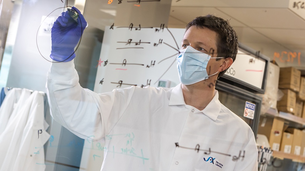 JAX assistant professor Ryan Tewhey writing on a board while wearing PPE